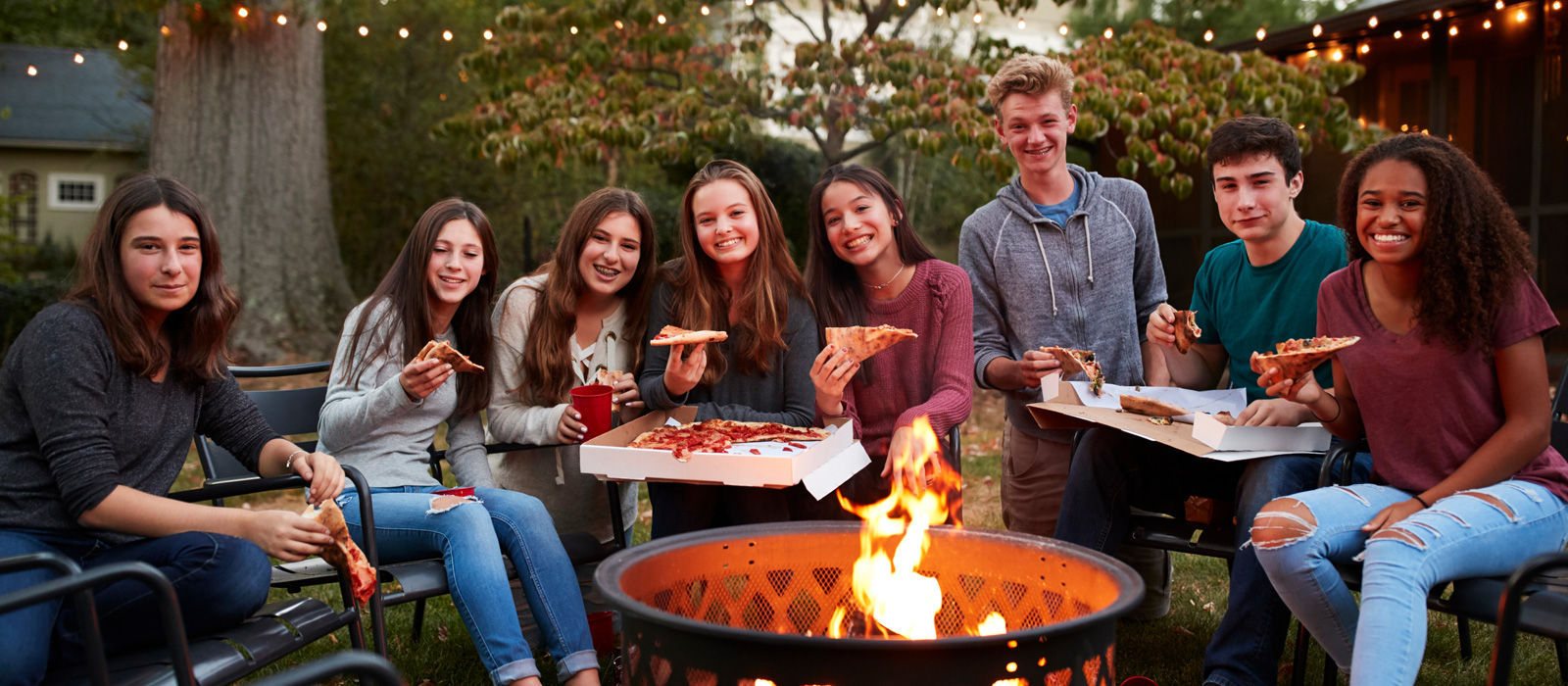 A group of high school aged students enjoying takeout pizza around a backyard bonfire